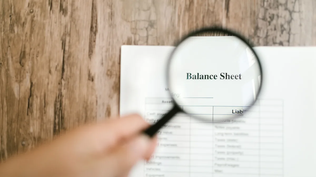 A hand holding a magnifying glass over a balance sheet document.